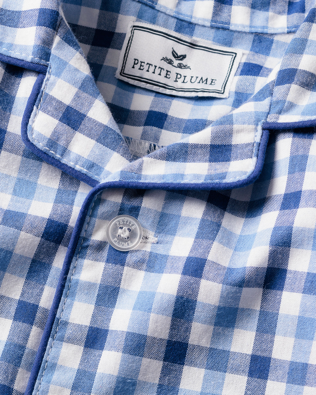 A close-up of the Babys Twill Summer Romper in Royal Blue Gingham by Petite Plume shows a collar with a fabric label, blue piping, and soft brushed fabric. A white button is fastened in the center.