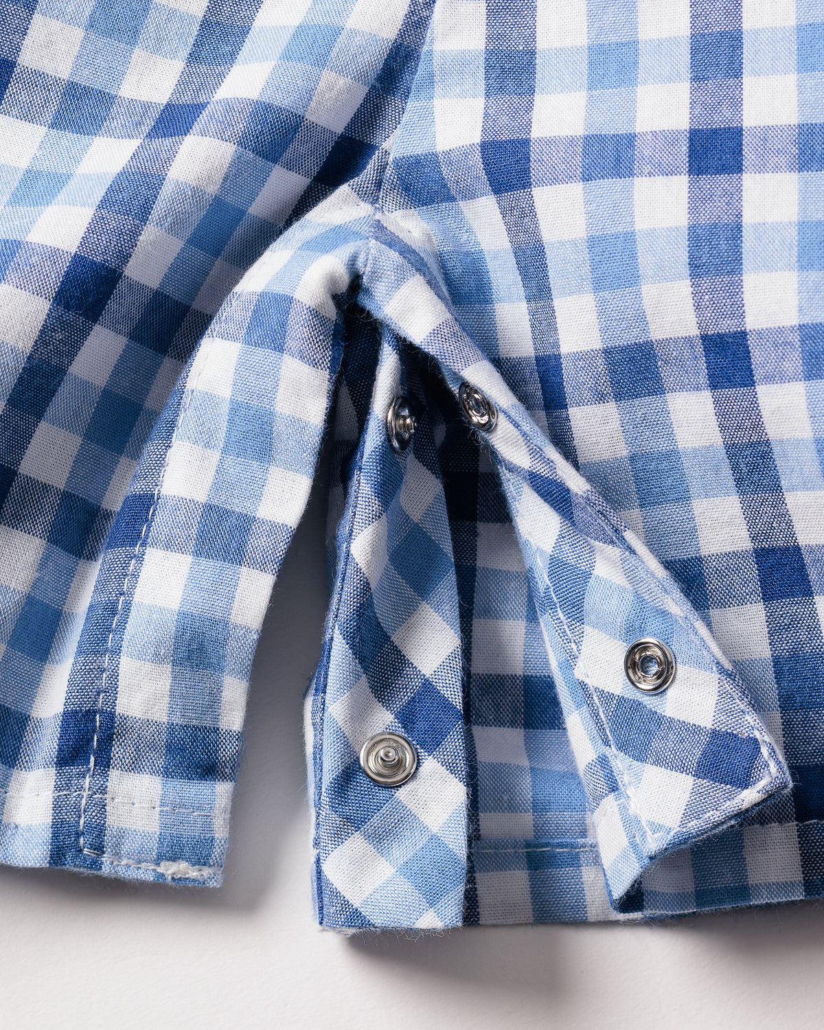 Close-up of Petite Plumes Babys Twill Summer Romper in Royal Blue Gingham, highlighting the metal snap buttons. The yarn-dyed cotton fabric is slightly folded to reveal detailed stitching and button features.