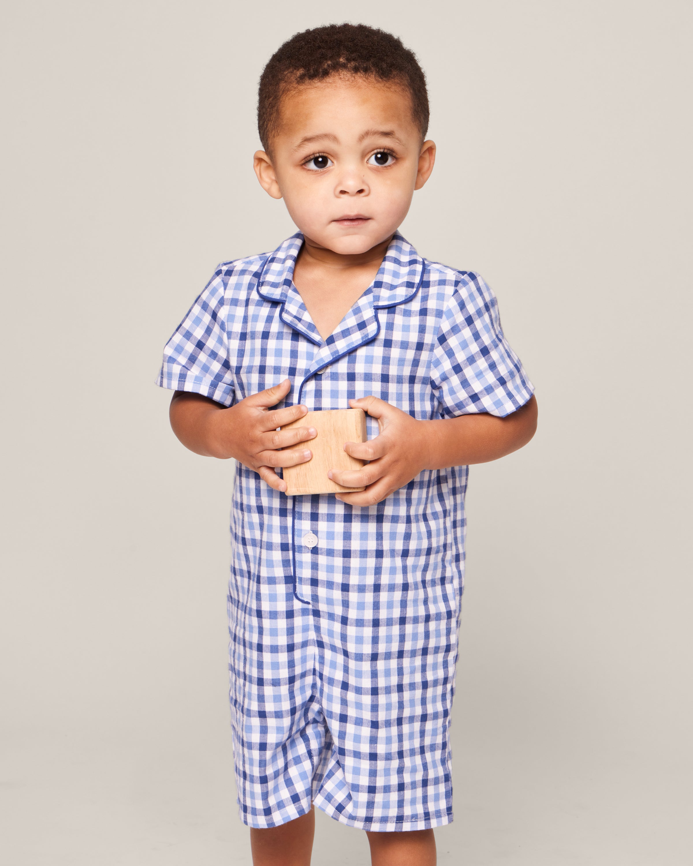 A toddler with short hair wears Petite Plumes Babys Twill Summer Romper in Royal Blue Gingham, holding a wooden block with both hands. The child looks off-camera with a neutral expression against a plain light gray background.
