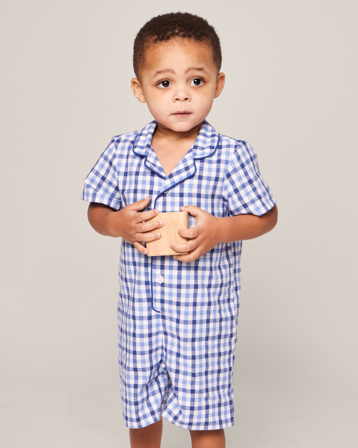 A toddler with short hair wears Petite Plumes Babys Twill Summer Romper in Royal Blue Gingham, holding a wooden block with both hands. The child looks off-camera with a neutral expression against a plain light gray background.