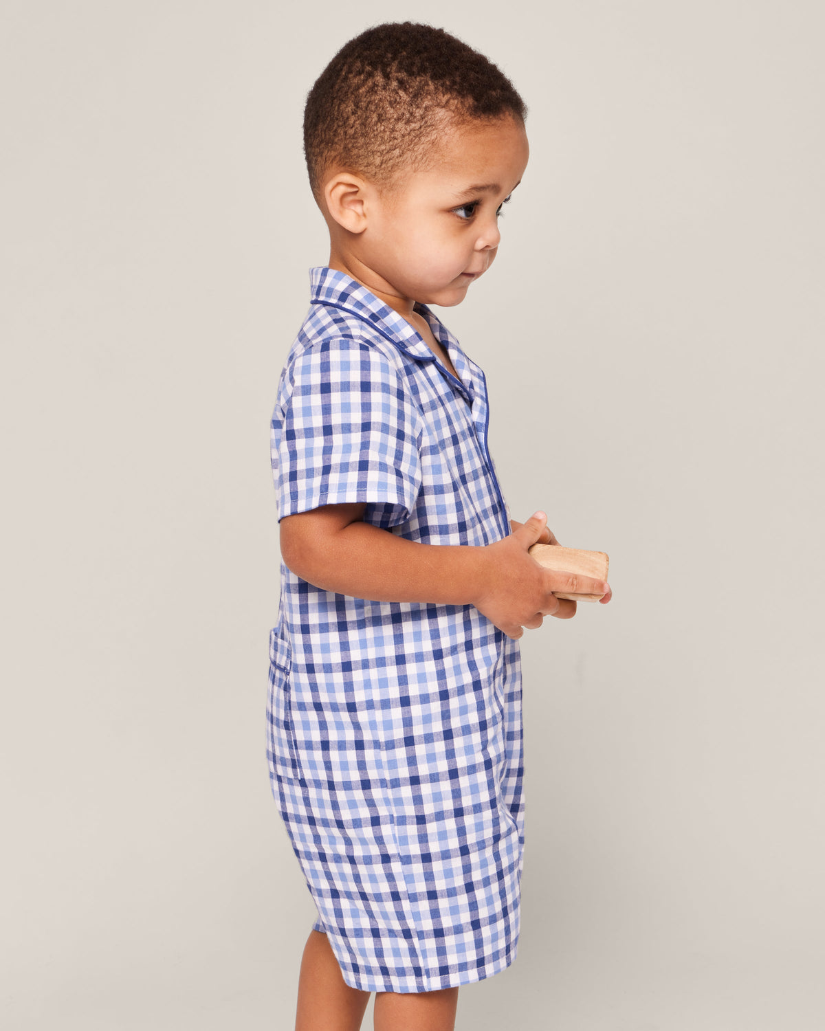 A young child stands in profile against a plain background, wearing Petite Plumes Babys Twill Summer Romper in Royal Blue Gingham, holding a light-colored wooden block, ready for imaginative play before slipping into sleepwear.