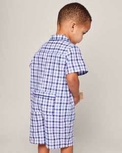 A young child stands facing away, clutching a small item. Dressed in Petite Plumes Babys Twill Summer Romper in Royal Blue Gingham with short sleeves and shorts, the plain background subtly emphasizes the charm of their outfit.