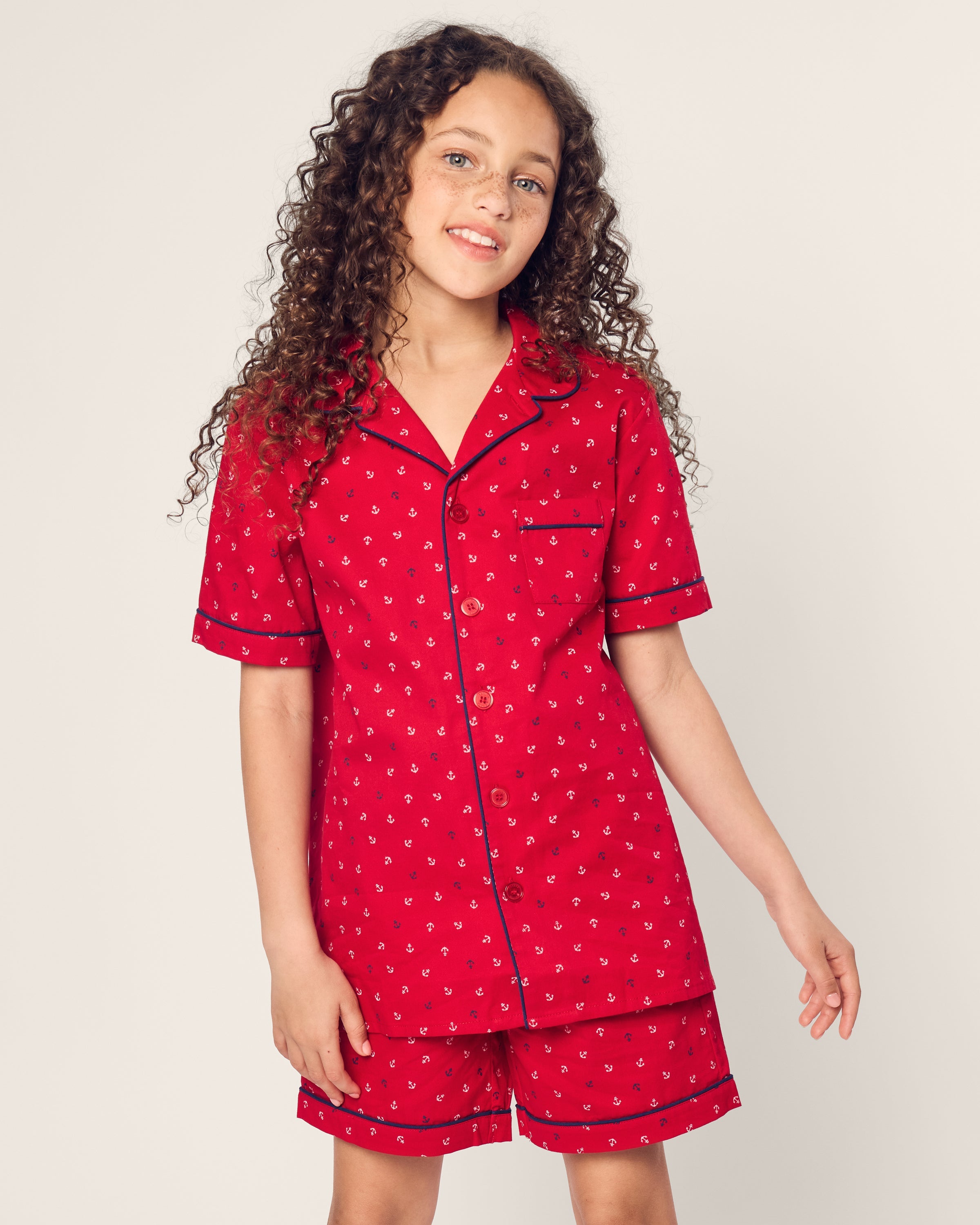A young girl with curly hair beams in her luxurious red Kids Twill Short Set in Anchors Away by Petite Plume. The flame-retardant ensemble features a short-sleeved, button-up top and matching shorts as she stands against a plain light background.