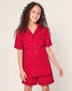 A young girl with curly hair beams in her luxurious red Kids Twill Short Set in Anchors Away by Petite Plume. The flame-retardant ensemble features a short-sleeved, button-up top and matching shorts as she stands against a plain light background.