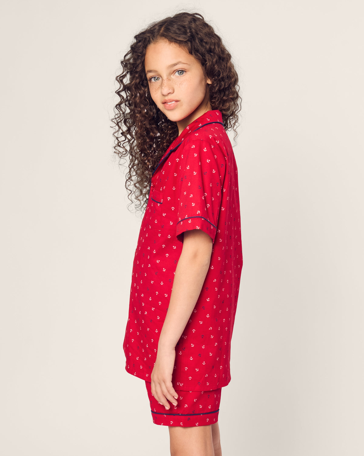 A young girl with long, curly hair stands sideways, gazing at the camera against a plain light background. She is wearing the Kids Twill Short Set in Anchors Away by Petite Plume.