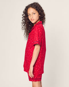 A young girl with long, curly hair stands sideways, gazing at the camera against a plain light background. She is wearing the Kids Twill Short Set in Anchors Away by Petite Plume.