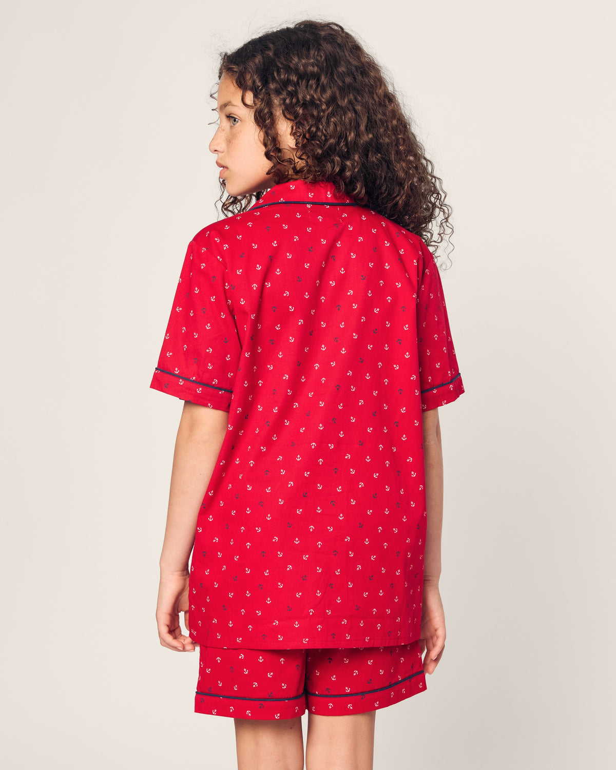 A child with curly hair, seen from the back, is wearing Petite Plumes red Kids Twill Short Set in Anchors Away, made from luxurious flame-retardant fiber. The pajamas feature a small print and black piping on an off-white background.