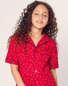A young person with long, curly hair gently smiles while wearing Petite Plumes Kids Twill Short Set in Anchors Away, featuring a short-sleeved, red button-up shirt made of flame retardant fiber with a small print design. The background is plain and light-colored.