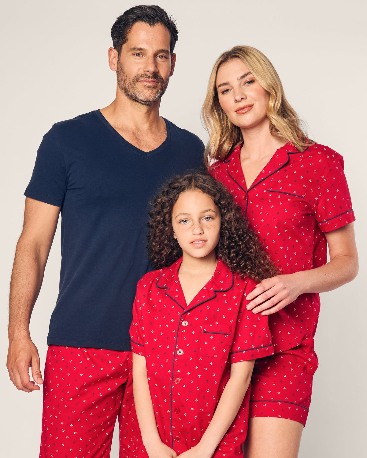 A man wears a navy blue T-shirt with red pajama bottoms, while a woman and child both wear matching luxurious red pajamas adorned with small patterns by Petite Plume. The childs set is the Kids Twill Short Set in Anchors Away. Crafted from flame retardant fibers, they ensure comfort and safety as they smile softly.