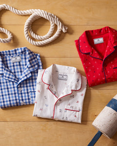 Three folded luxurious pajama tops by Petite Plume lie on a nautical-themed wooden surface. The tops include a Kids Twill Short Set in Anchors Away, with blue gingham, white with small nautical prints, and red with tiny anchor designs. A coiled rope and buoy complete the display.