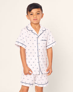 A young boy wears Petite Plumes Kids Twill Short Sleeve Short Set in Buzzing Bees, featuring white pajamas with buzz bee print and navy piping. The set has a button-up top and matching shorts, exuding classic comfort and style against a light background.