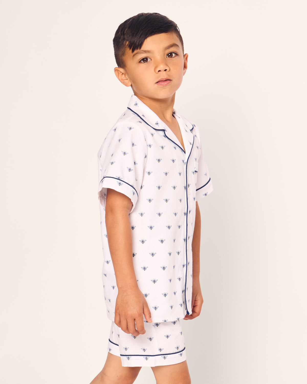 A young boy with short dark hair wears Petite Plumes Kids Twill Short Sleeve Short Set in Buzzing Bees, featuring luxurious flame-retardant fabric with a small pattern and dark piping. The pajama set has a short-sleeved, button-up shirt, and he gazes slightly upward against a plain background.