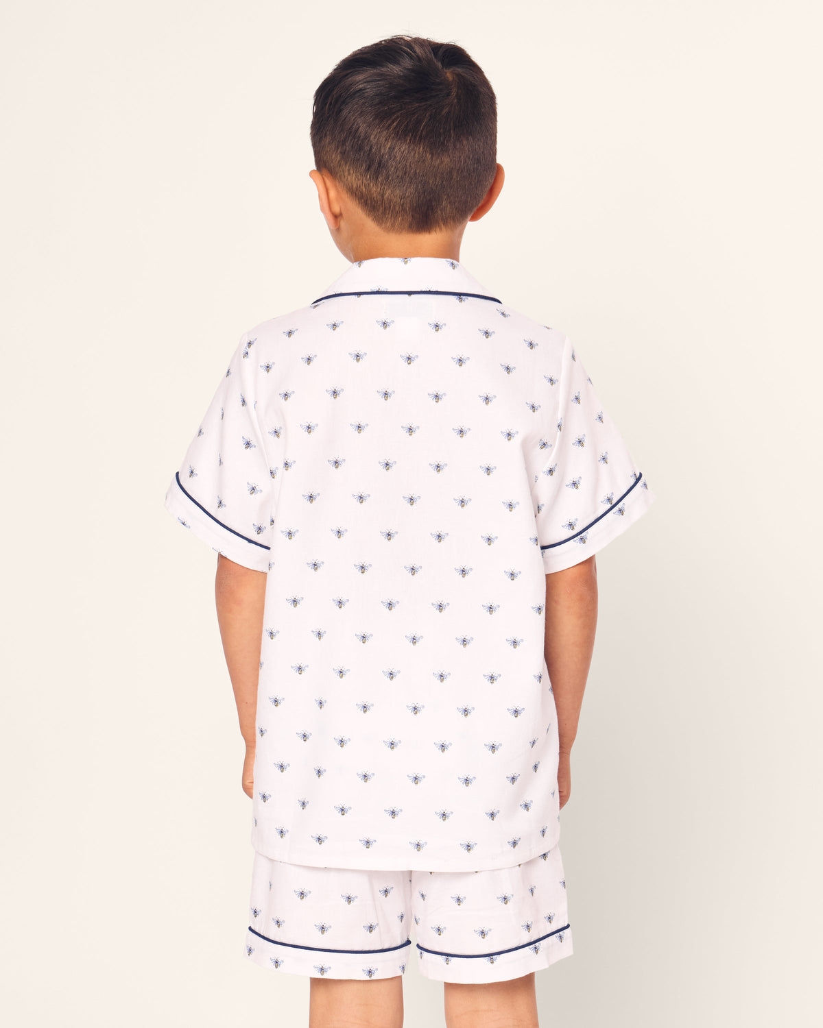 A young boy, seen from the back, wears Petite Plumes luxurious Kids Twill Short Sleeve Short Set in Buzzing Bees print. These white pajamas feature small blue bees and short sleeves accented with blue piping.