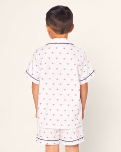 A young boy, seen from the back, wears Petite Plumes luxurious Kids Twill Short Sleeve Short Set in Buzzing Bees print. These white pajamas feature small blue bees and short sleeves accented with blue piping.