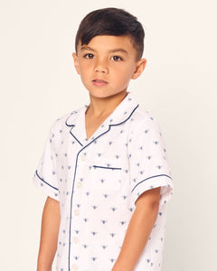 A young boy wears the Kids Twill Short Sleeve Short Set in Buzzing Bees by Petite Plume, featuring white pajamas with blue bee patterns and black piping. His short dark hair is slightly tousled as he looks to the side against a plain off-white background.