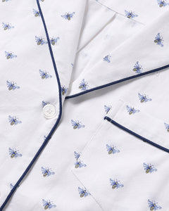 An up-close view of a white fabric with a Buzzing Bees print from Petite Plume showcases small blue and yellow bees, alongside detailed elements like a button, blue piped lapel, and pocket. This suggests luxurious sleepwear such as the Kids Twill Short Sleeve Short Set in Buzzing Bees.