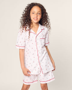 A young girl with curly hair wears the Petite Plume Kids Twill Short Sleeve Short Set in Coastal Charms, featuring white pajamas with red piping. She stands against an off-white background, slightly smiling with her hands by her side.