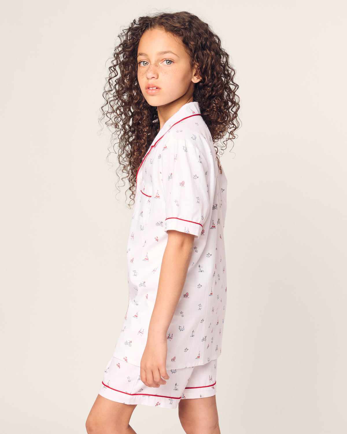 A young girl with curly hair wears the Petite Plume Kids Twill Short Sleeve Short Set in Coastal Charms—white pajamas with red trim and small colorful patterns. She gazes to the side against a plain background, exuding comfort and style.