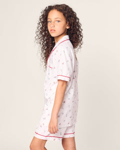 A young girl with curly hair wears the Petite Plume Kids Twill Short Sleeve Short Set in Coastal Charms—white pajamas with red trim and small colorful patterns. She gazes to the side against a plain background, exuding comfort and style.