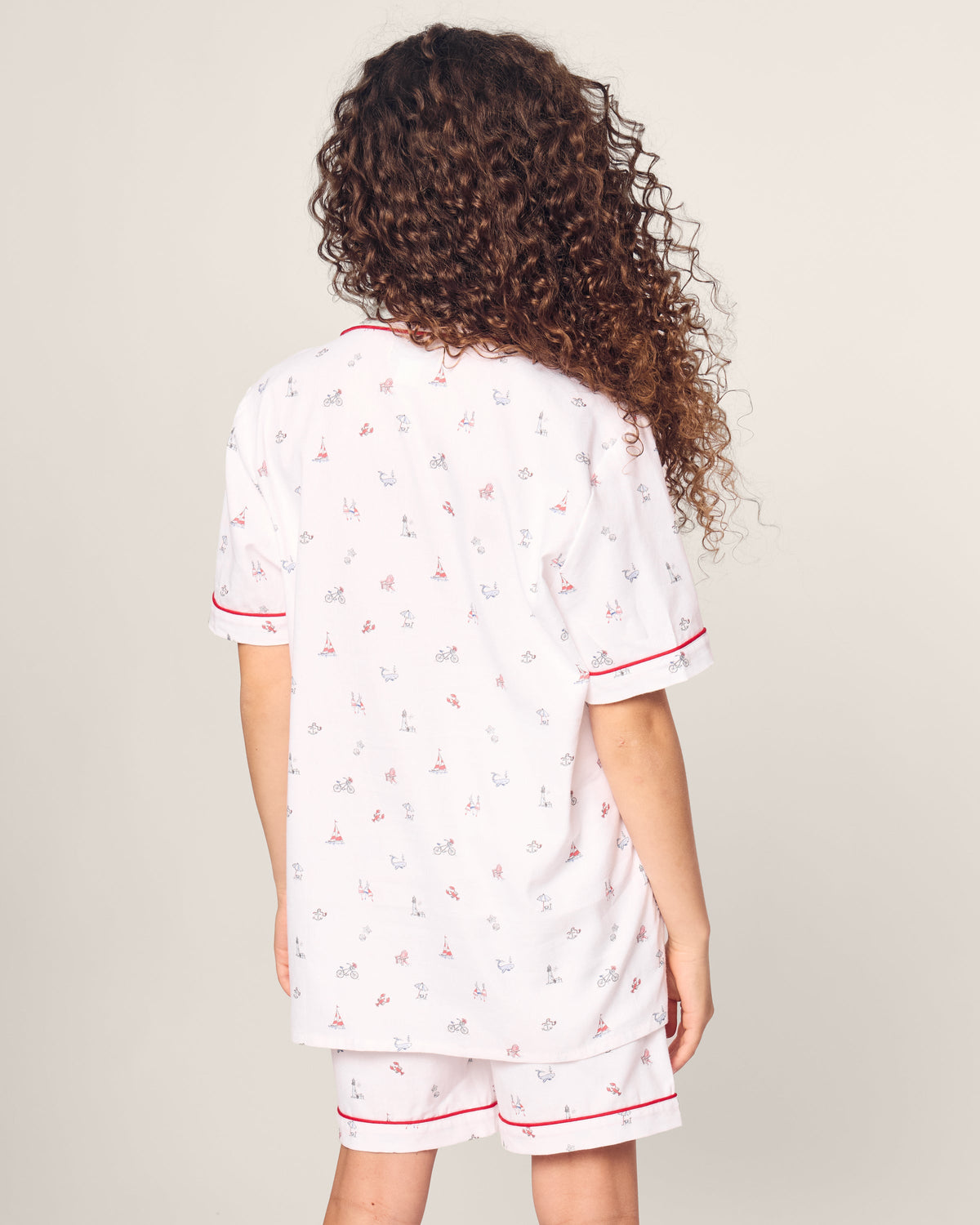 A child with long curly hair wears Petite Plumes Kids Twill Short Sleeve Short Set in Coastal Charms, featuring white pajamas with red trim. They stand facing away, hands by their sides against a plain light gray background.