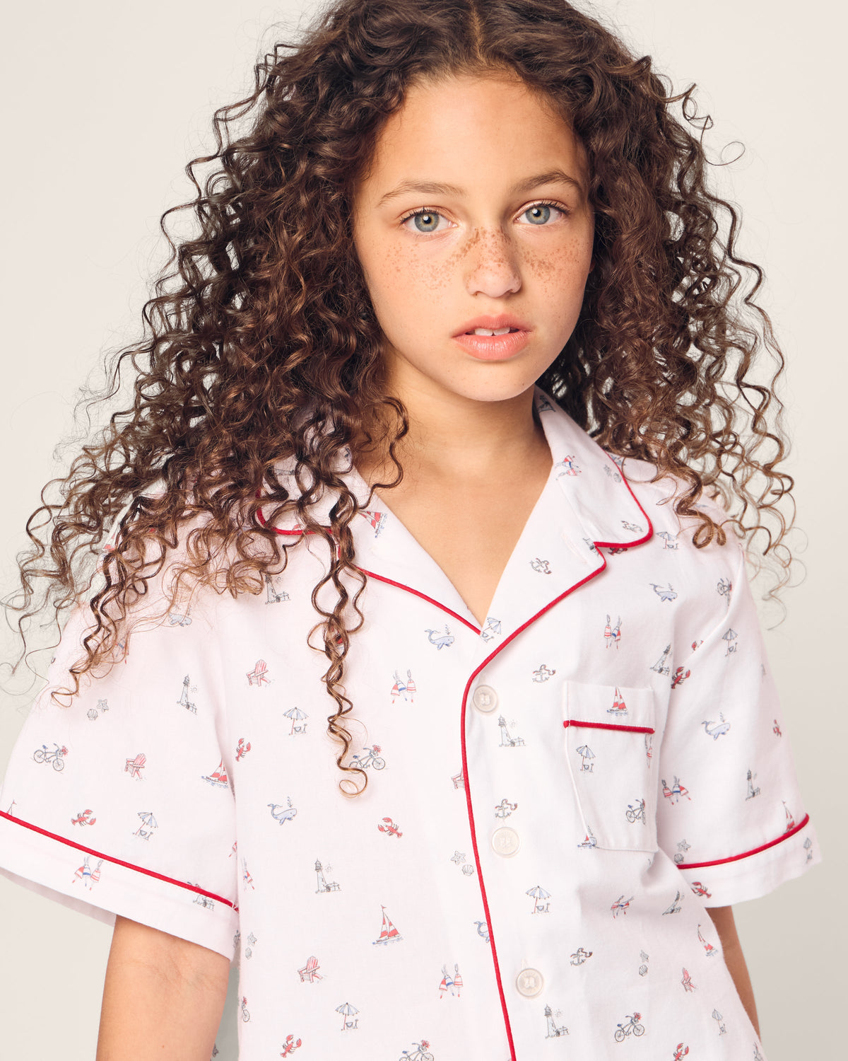 A young girl with curly hair is wearing Petite Plumes Kids Twill Short Sleeve Short Set in Coastal Charms, featuring lively prints and red trim, as she looks at the camera. Neutral background.