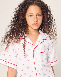 A young girl with curly hair is wearing Petite Plumes Kids Twill Short Sleeve Short Set in Coastal Charms, featuring lively prints and red trim, as she looks at the camera. Neutral background.
