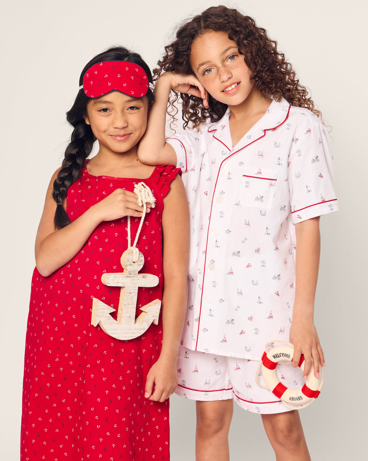 Two smiling girls in flame-retardant sleepwear, one in a red nightdress with anchor decor and sleep mask, the other in Petite Plumes Coastal Charms Kids Twill Short Sleeve Short Set with nautical patterns. They hold a small lifebuoy saying WELCOME ONBOARD.