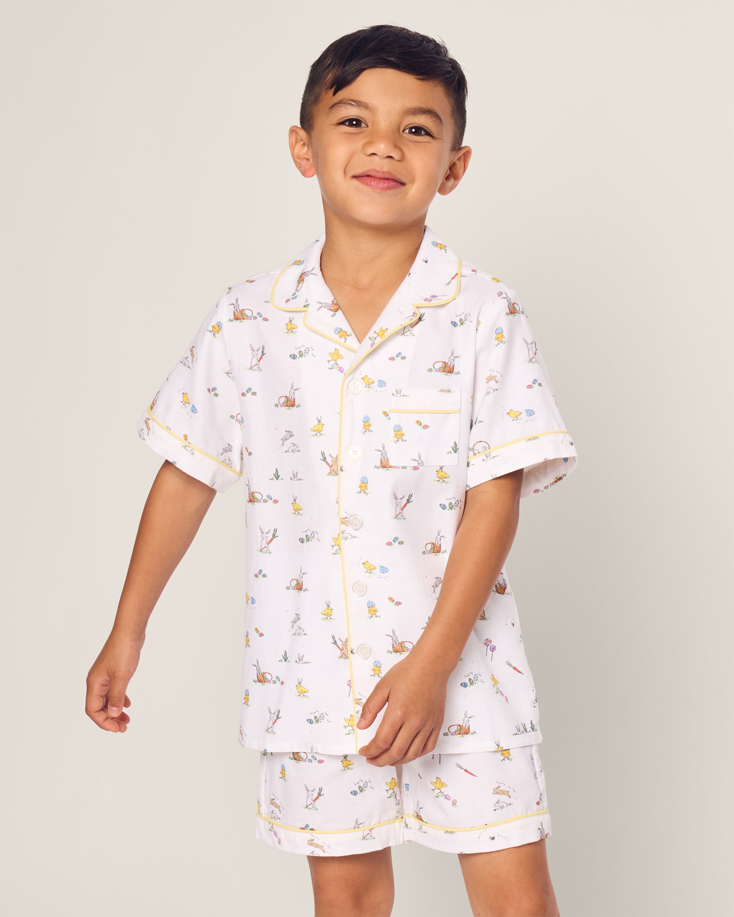 A young child beams in Petite Plumes Kids Twill Short Set in Easter Frolic, the light-colored, short-sleeved luxurious pajamas feature playful prints. The plain background highlights their cheerful expression and adorable pajama design.