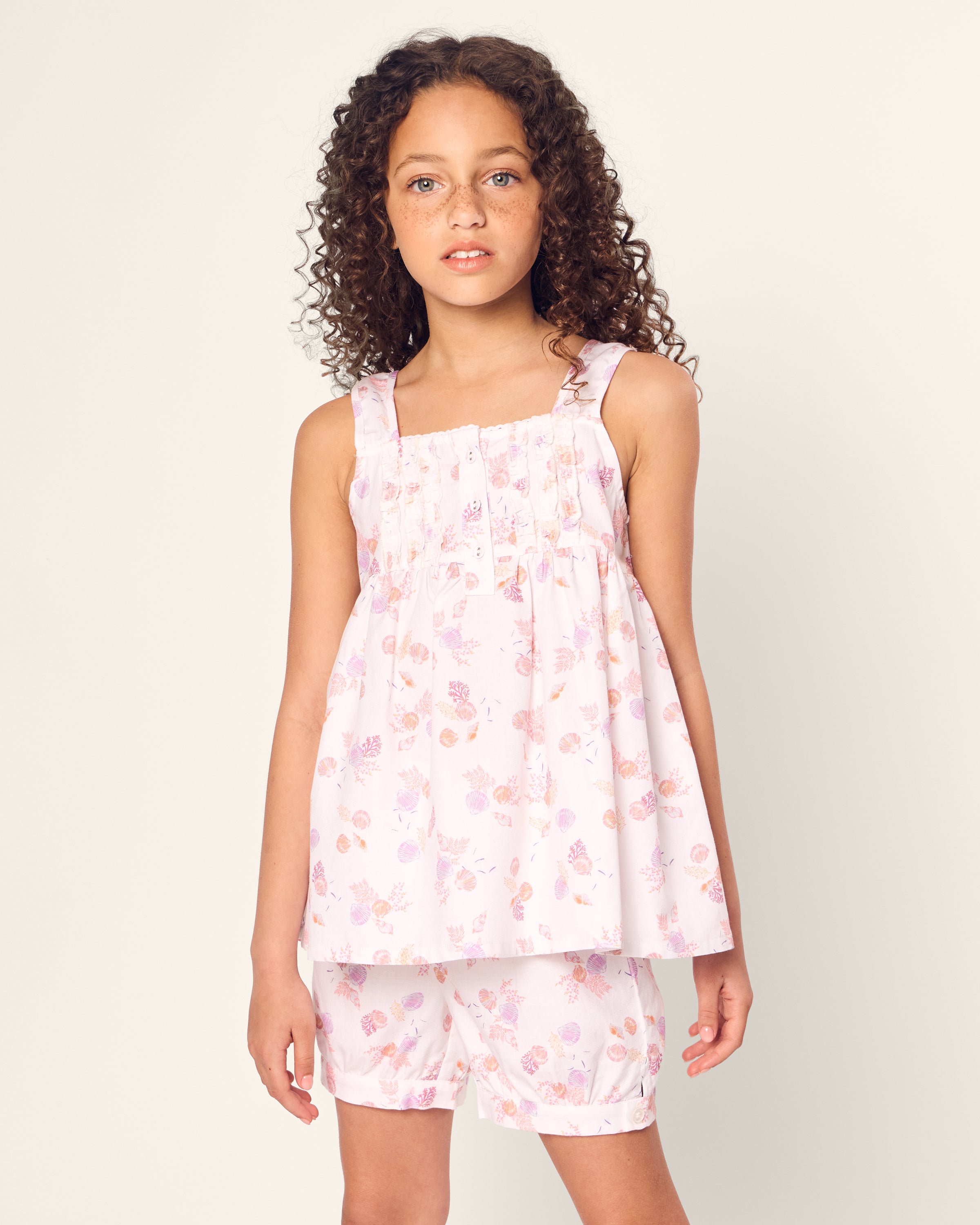 A young girl with curly hair stands against a plain light background, wearing the Girls Twill Charlotte Short Set in Seashell Splendor by Petite Plume. Her sleeveless top and matching shorts exude luxurious sleepwear vibes as she gazes toward the camera with a neutral expression.