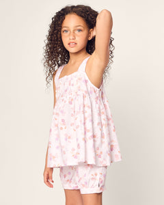A young girl with long curly hair poses with one arm raised, in her stylish Petite Plume Girls Twill Charlotte Short Set in Seashell Splendor, showing off the light pink floral sleeveless dress and matching shorts against a plain light background.