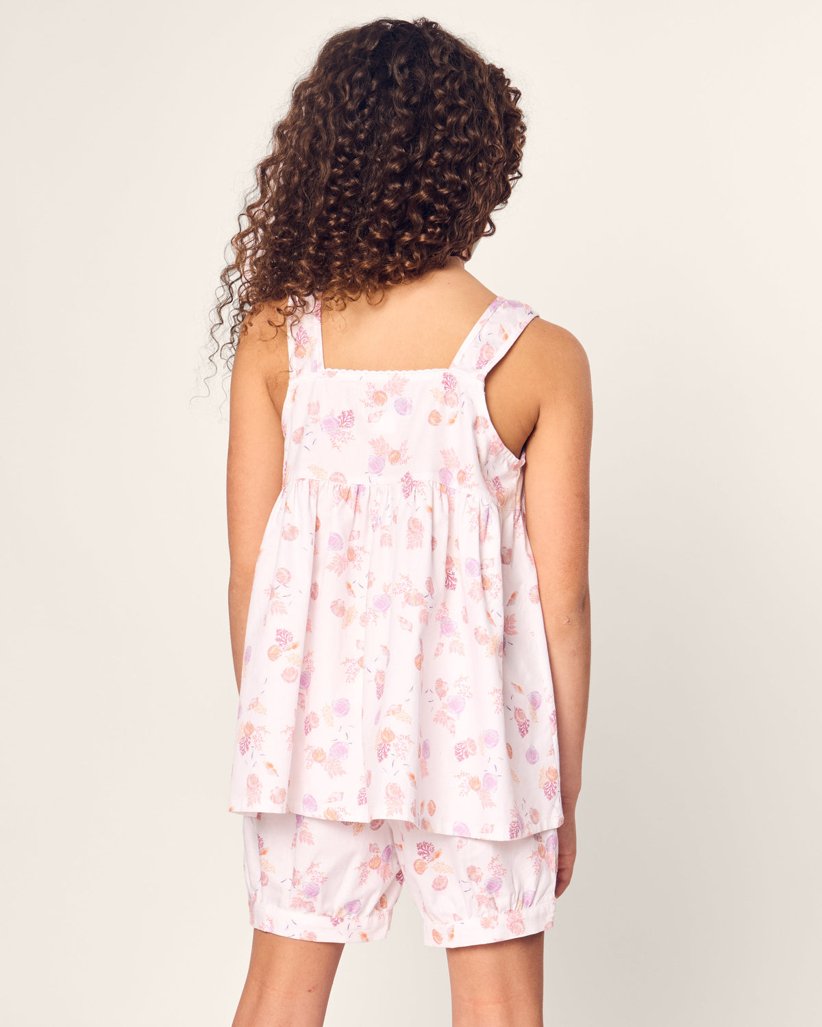 A young girl with curly hair stands facing away, wearing Petite Plumes Twill Charlotte Short Set in Seashell Splendor, featuring a pink floral sleeveless top and matching shorts made from flame retardant fiber, set against a plain background.