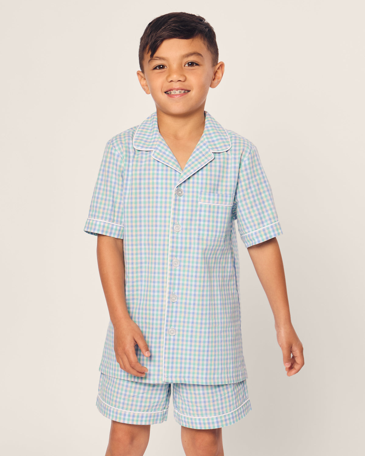 A young boy beams in his Petite Plume Kids Twill Short Set, a luxurious cotton sleepwear with short sleeves and button-up design. The charming blue and green Spring Gingham print stands out against the plain, light-colored background.