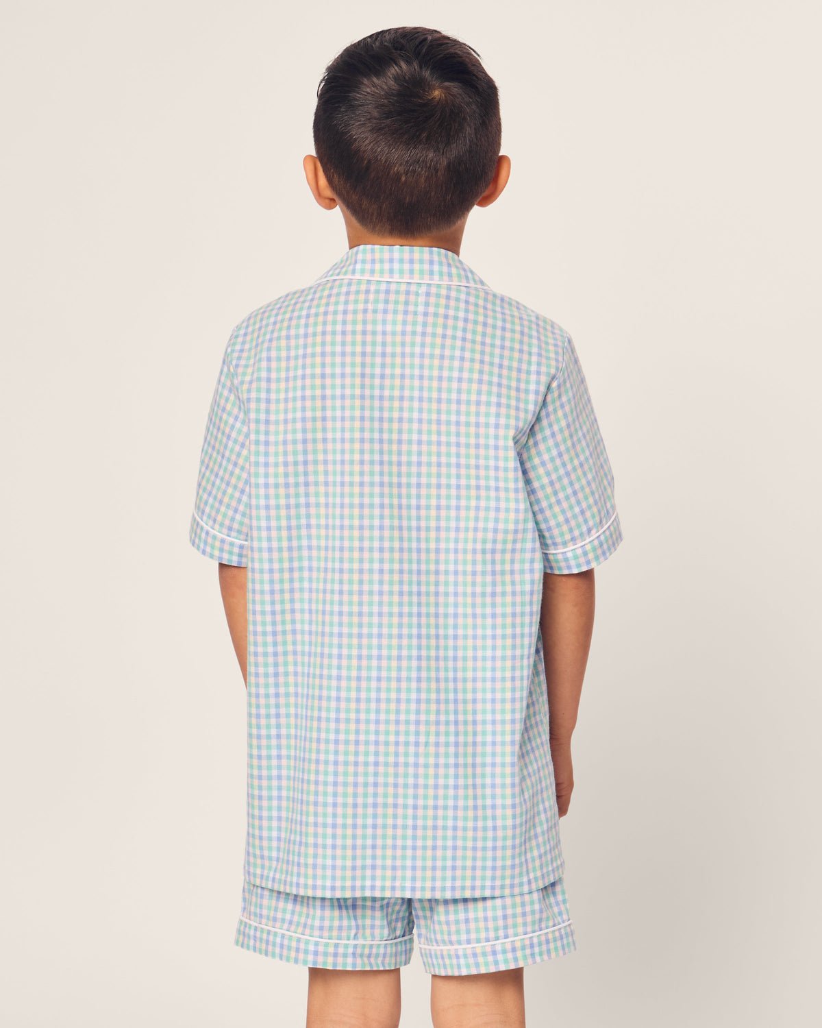 A young boy is seen from behind, wearing Petite Plumes Kids Twill Short Set in Spring Gingham with pastel blue and light purple checks, standing against a white background.