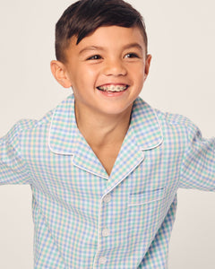 A smiling child in a Petite Plume Kids Twill Short Set in Spring Gingham showcases a light blue and green gingham print pajama top with white piping and buttons. Their short dark hair adds to the look as they gaze sideways against a plain background.