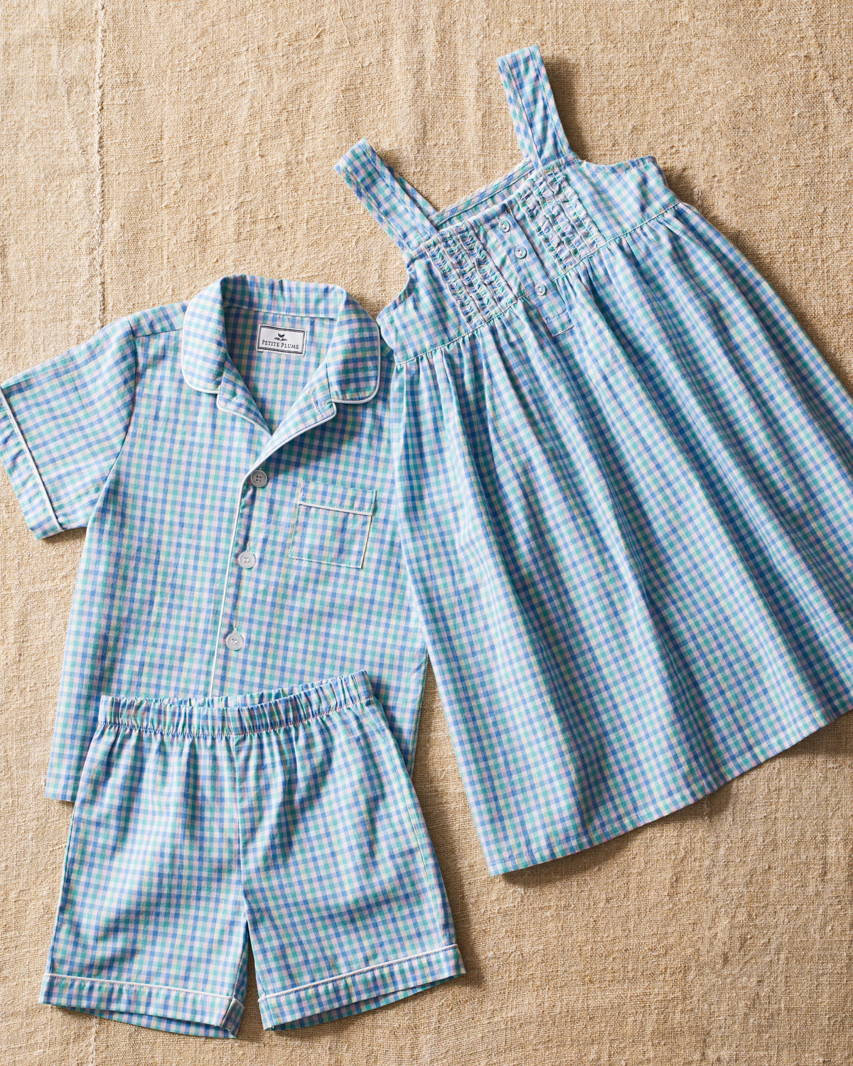 The Petite Plume Kids Twill Short Set in Spring Gingham, displayed on a textured beige surface, features luxurious cotton sleepwear with a blue and green gingham print. It includes a short-sleeve button-up shirt, matching shorts, and sleeveless nightgown in the same charming pattern.