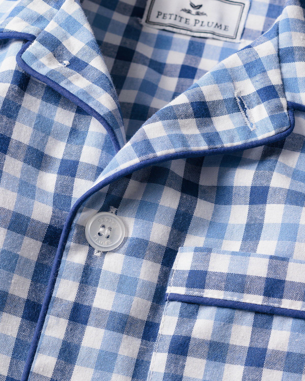 Close-up of a blue and white gingham patterned shirt from the Kids Twill Pajama Short Set in Royal Blue Gingham. It includes buttons, a pocket with piping detail, reflecting luxurious sleepwear charm. The neck label reads Petite Plume.