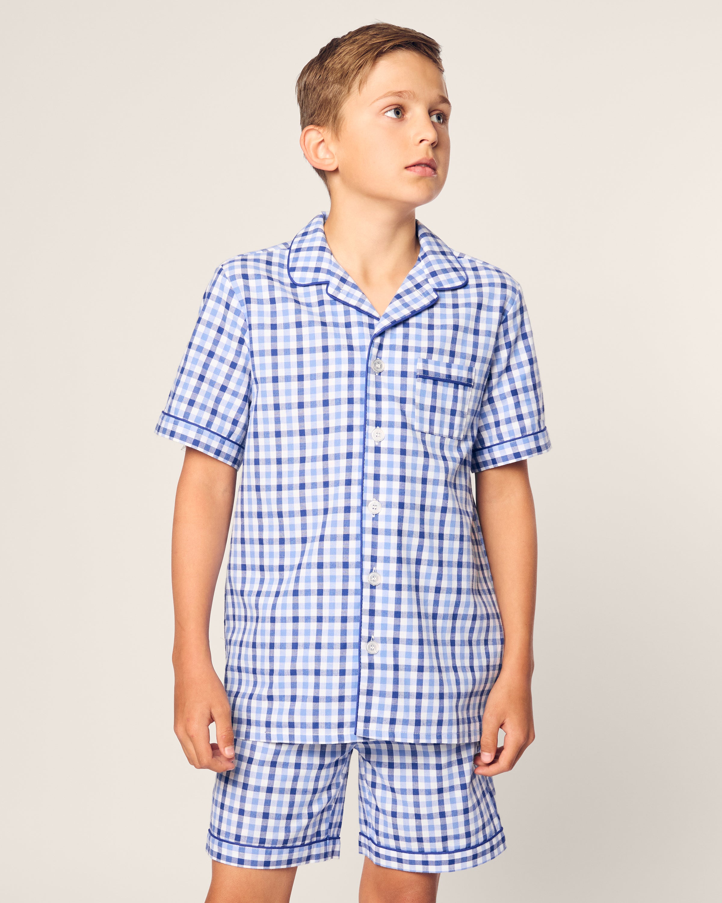 A young boy models the Kids Twill Pajama Short Set in Royal Blue Gingham by Petite Plume. The set features a yarn-dyed cotton short-sleeved button-up shirt with a collar and chest pocket, paired with shorts, as he gazes to the side against a plain backdrop.