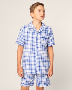 A young boy models the Kids Twill Pajama Short Set in Royal Blue Gingham by Petite Plume. The set features a yarn-dyed cotton short-sleeved button-up shirt with a collar and chest pocket, paired with shorts, as he gazes to the side against a plain backdrop.