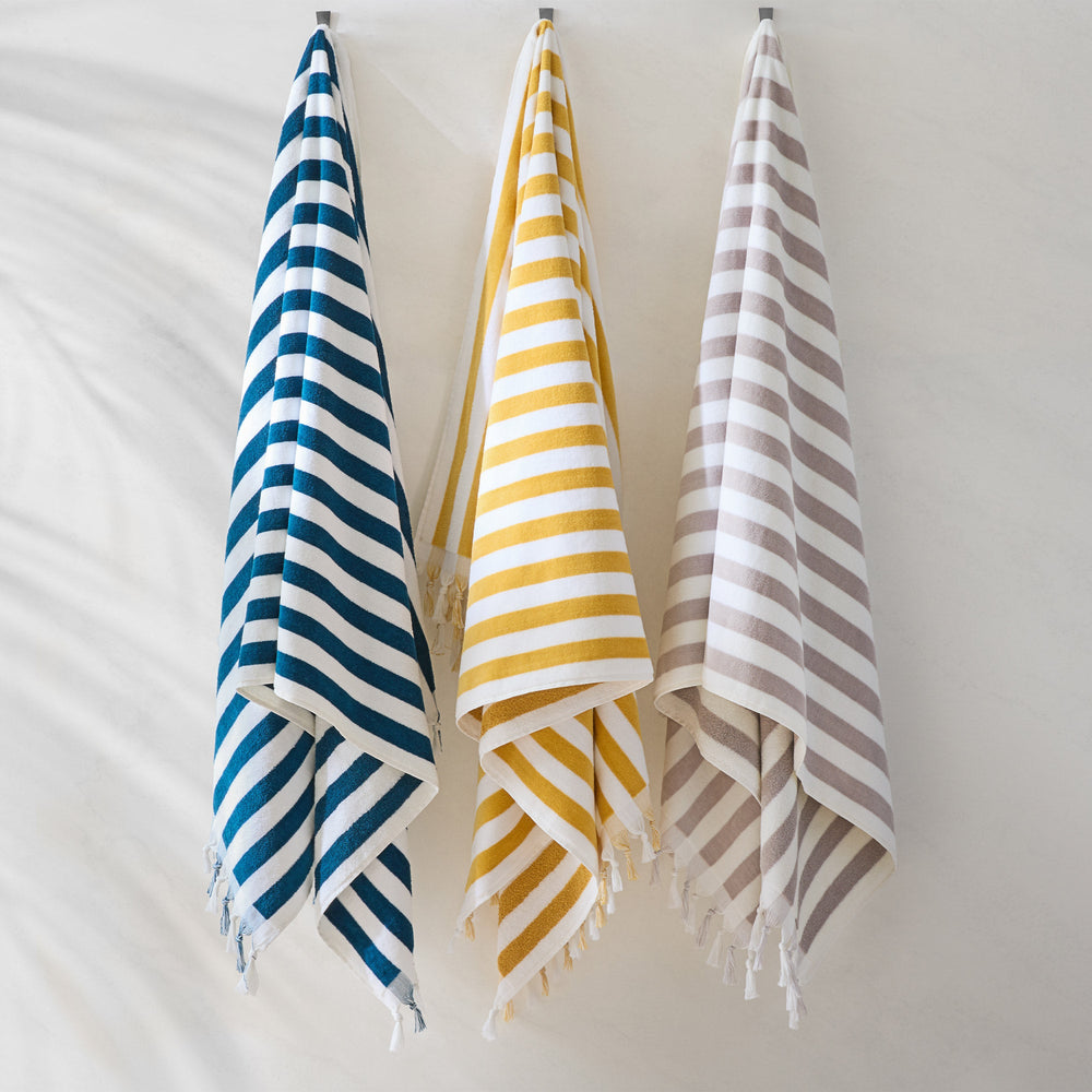 Front view of Cannes beach towels showcasing navy/white, yellow/white, and grey/white stripes with fringe against a light wall