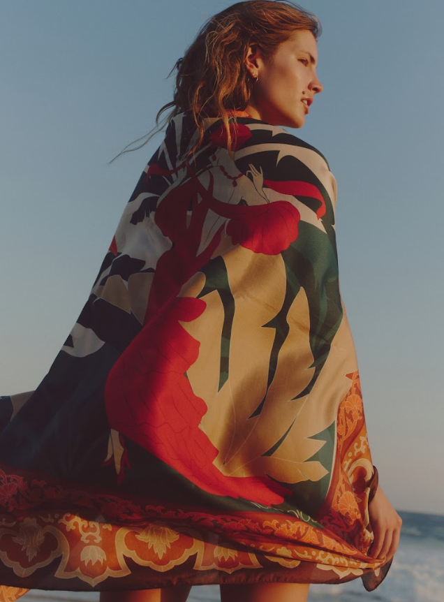 Back view of De La Falaise cotton-silk sarong showcasing bold floral and ornamental border print draped on model at the beach