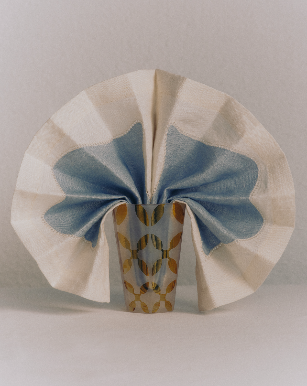 Close view of Les Courbes Napkin in Celeste showcasing its blue wave motif draped around a patterned glass on a neutral background