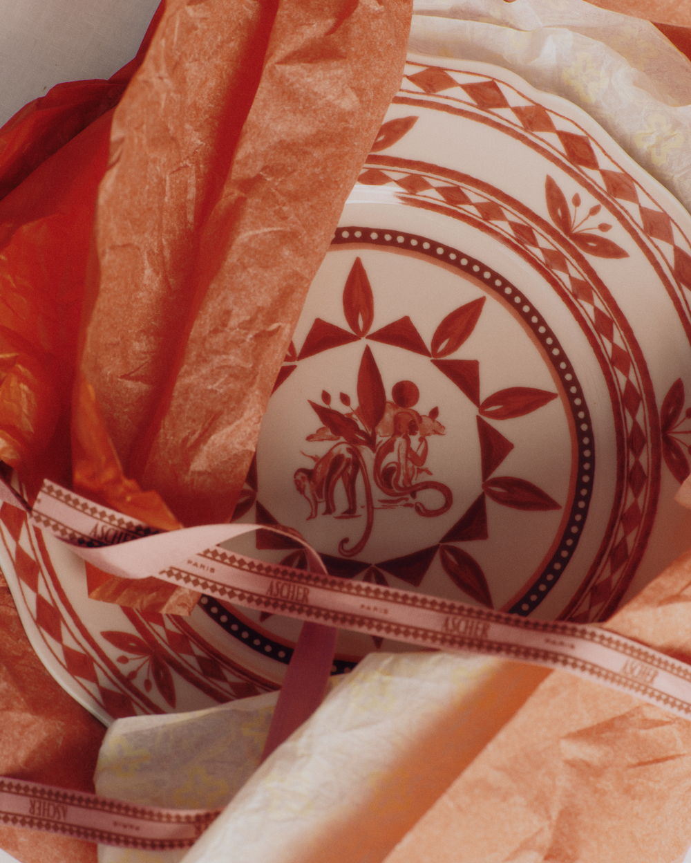 Close-up top view of Rêverie des Pins dining plate in Carmin showcasing red gouache botanical and monkey illustration on white porcelain, nestled in orange tissue and ribbon