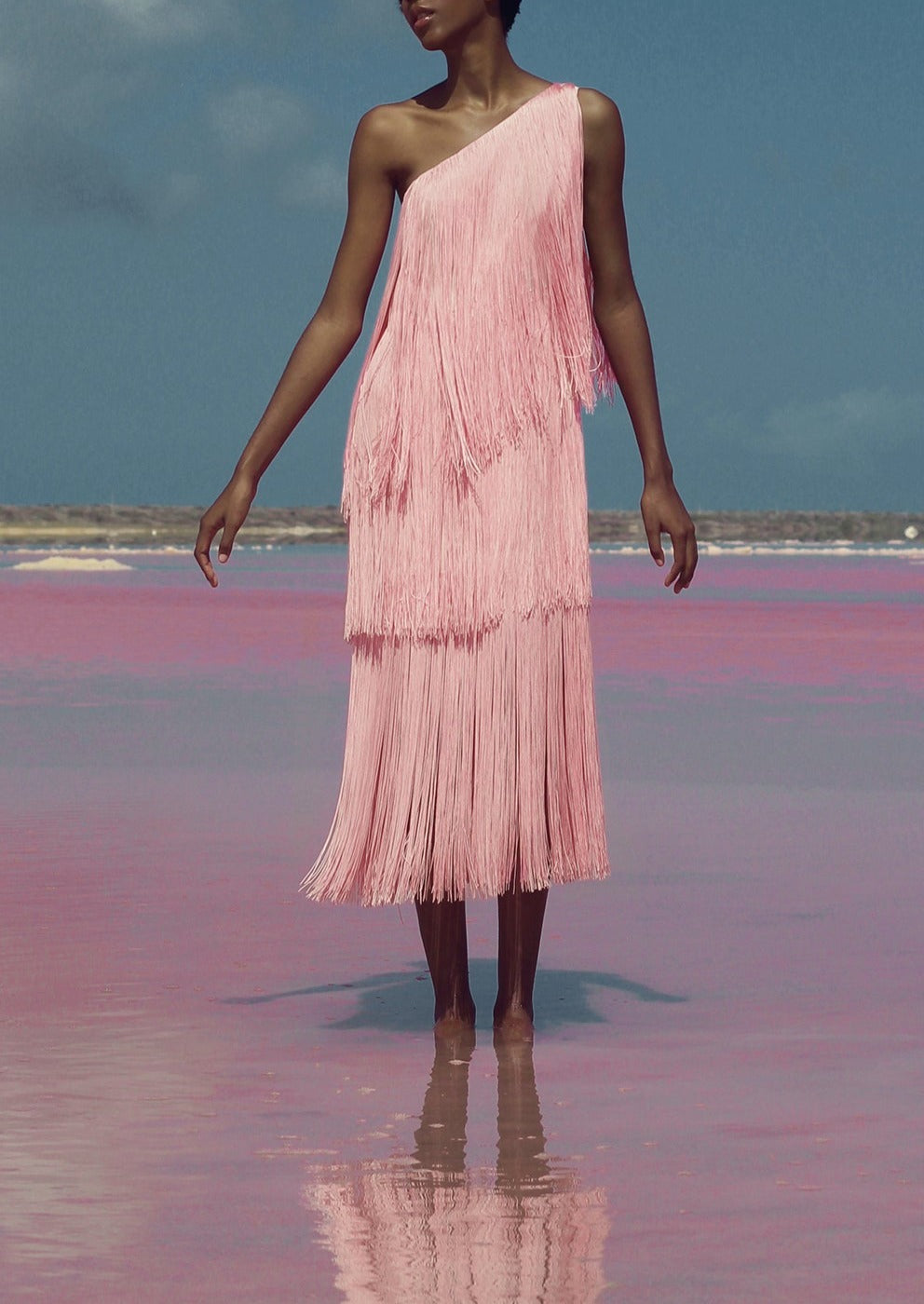 Front view of the Cascada Dress in Pink showcasing its one-shoulder neckline and layered fringe on a model standing in shallow water