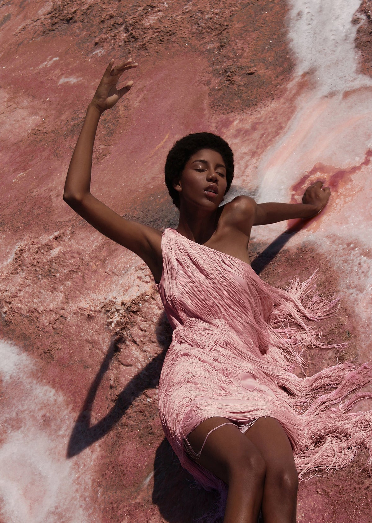 Full-length view of Cascada Dress In Pink showcasing one-shoulder styling and layered fringe on a model against a pink textured background