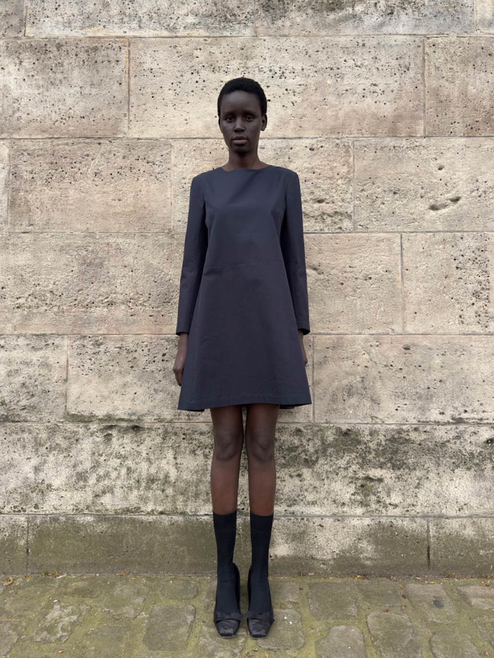 Front view of Catherine Dress in cotton gabardine on a model; long sleeves, A-line, above-knee length, stone wall background