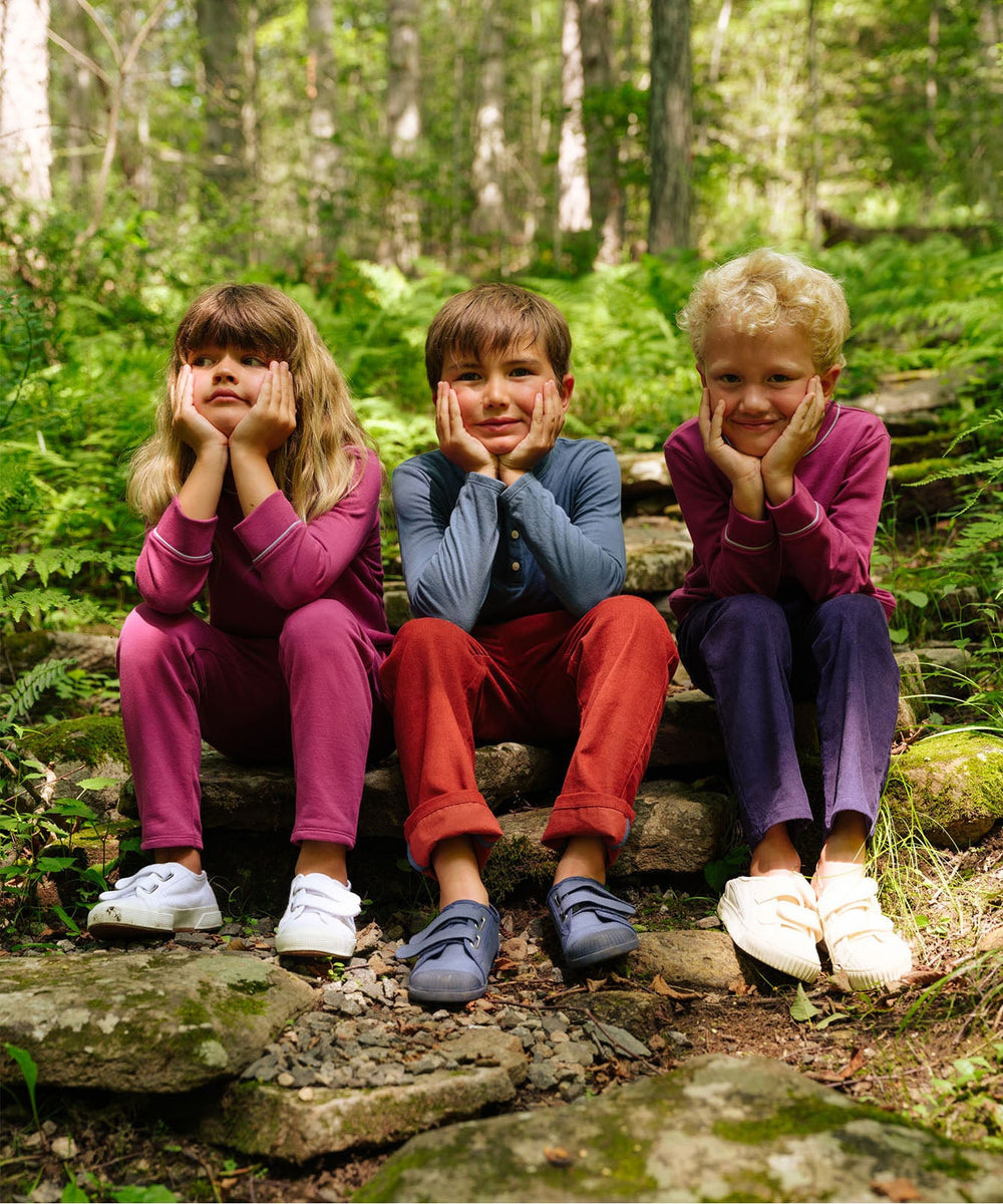Three children sitting on stone steps in a forest, leaning on their hands and wearing Chris Jogger pants including the blackberry color