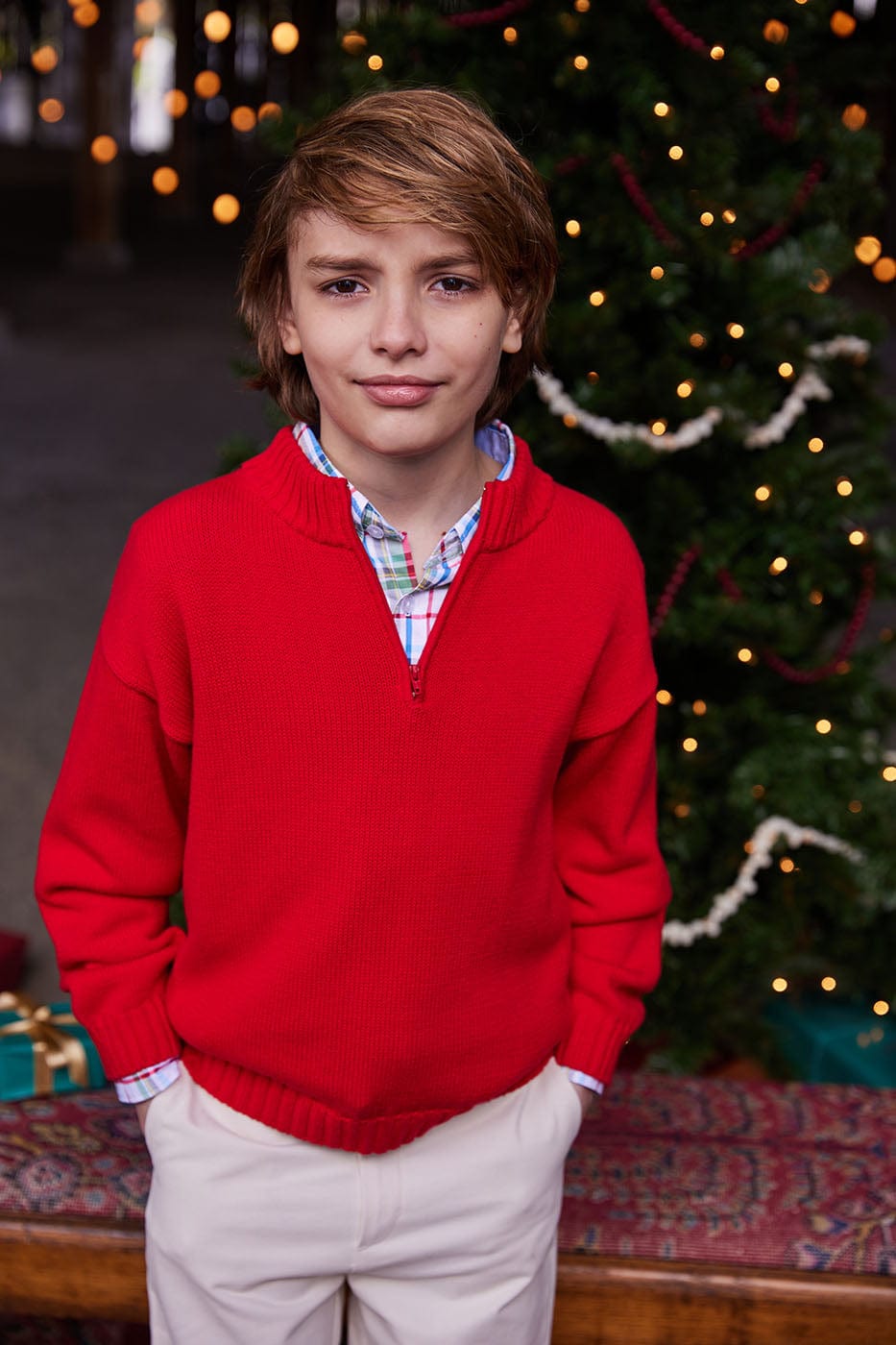Boy in red quarter-zip sweater over plaid shirt standing by decorated Christmas tree
