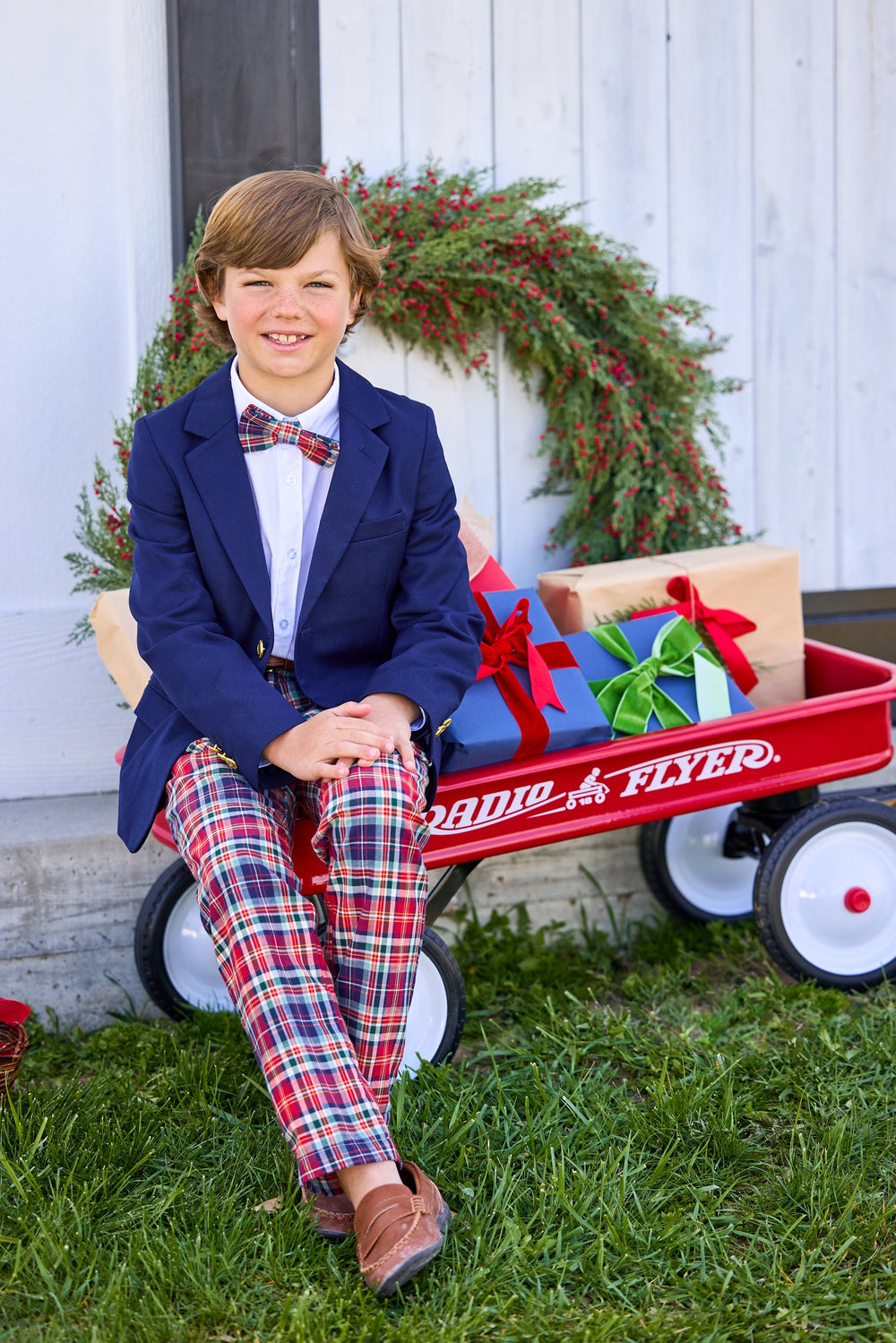 Front view of Classic Pant in Middleburg Plaid on a boy, red navy and hunter green plaid slim fit, seated by a red wagon