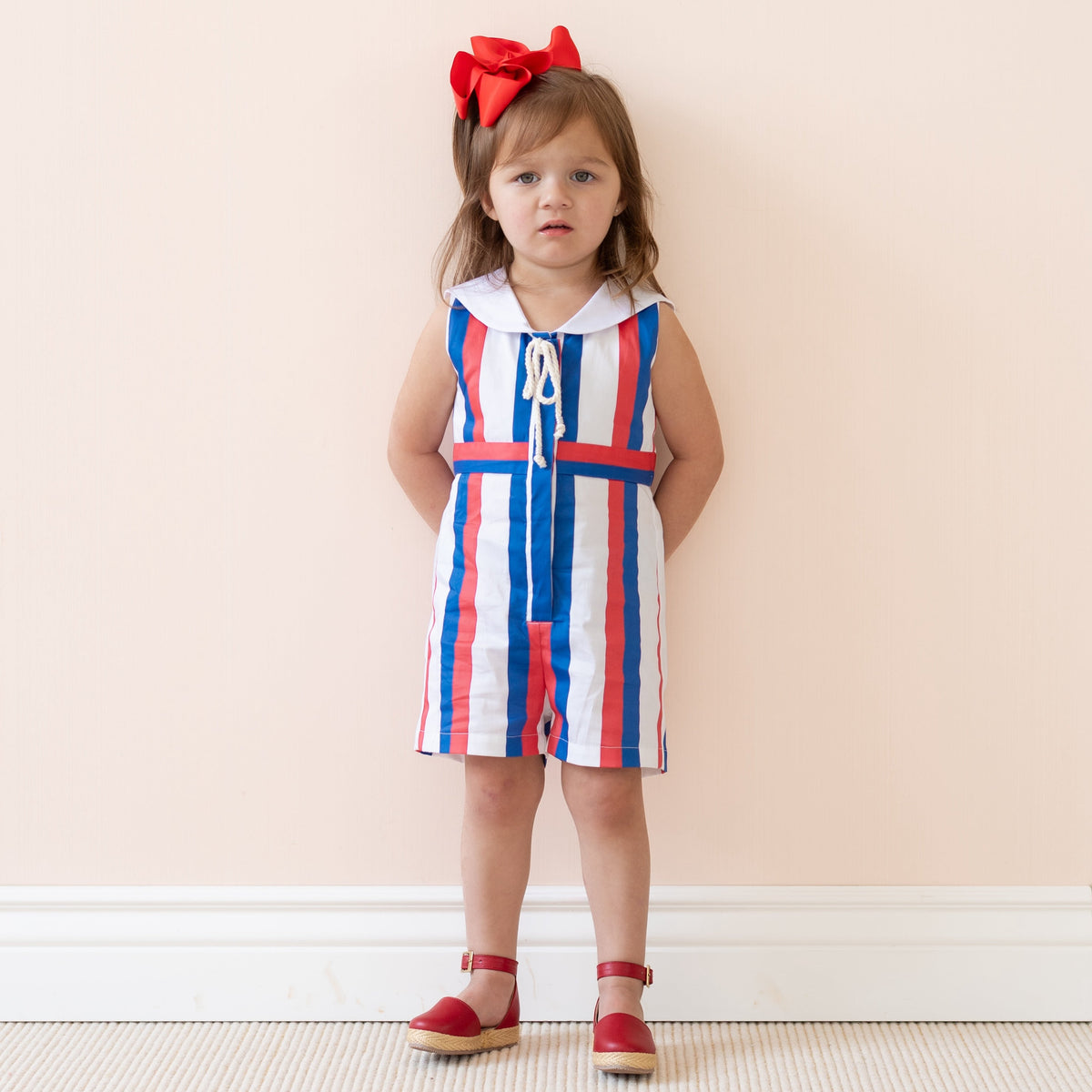 Front view of Sarah Girl Romper on child model showing sailor collar, nautical rope bow, and red, white, and blue stripes