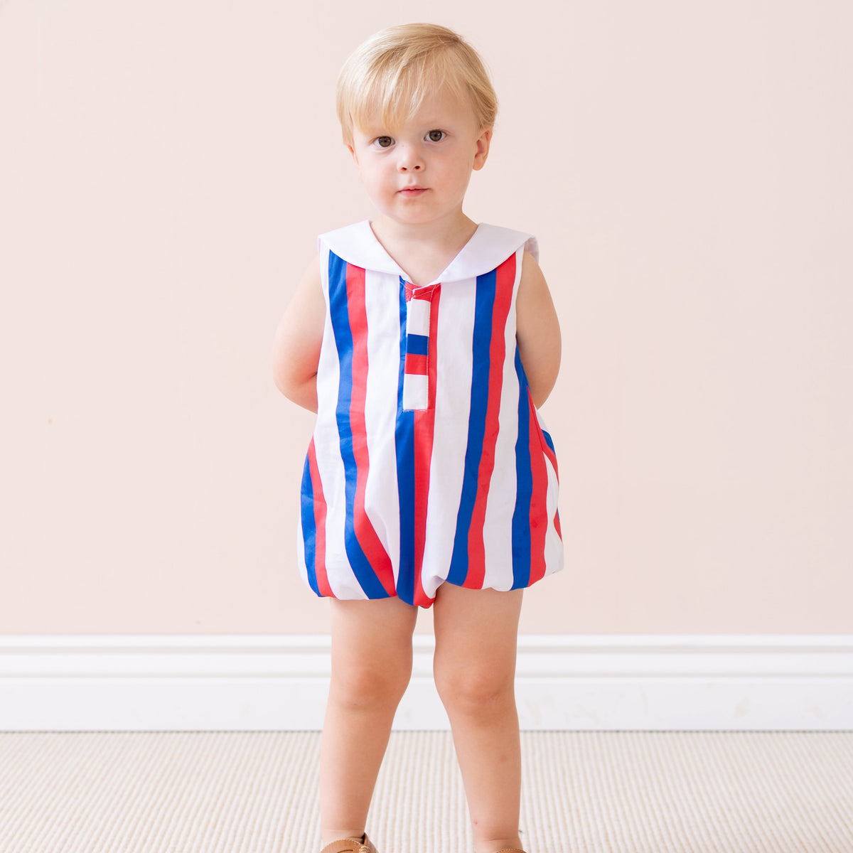 Front view of Sam Boy Bubble romper on a toddler with red, white and blue vertical stripes, white sailor collar, and bubble fit against a pale pink background